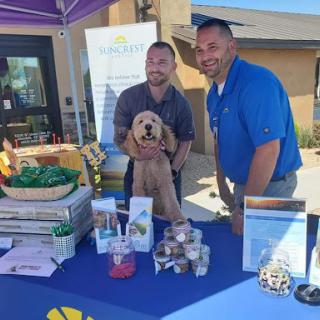 Two smiling individuals posing with a dog at an outdoor event booth.