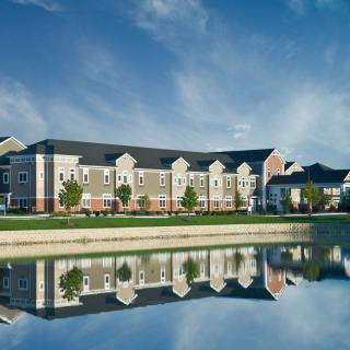 A residential building reflected in a calm lake with a clear blue sky and scattered clouds.