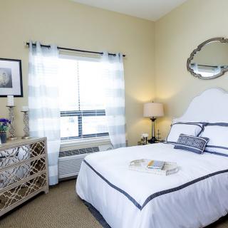 A cozy and minimalist bedroom with neutral tones, featuring a neatly made bed, a lamp on the bedside table, and light curtains covering the window.