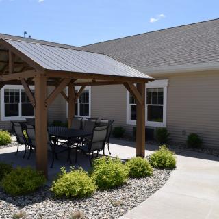 A sunny patio area features a wooden pergola with outdoor furniture, surrounded by landscaping.
