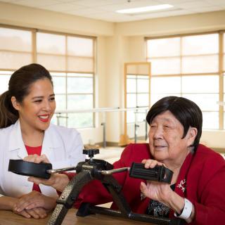 A healthcare professional assists an elderly woman with physical therapy using exercise equipment.