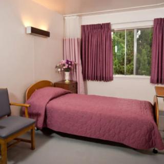 A neatly arranged bedroom featuring a single bed with pink bedding, pink curtains, a window view, a wooden desk and chair, and a wall-mounted lamp.