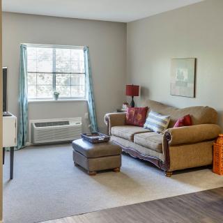 A cozy living room with a plush sofa and ottoman, decorated with cushions and artwork, featuring a large window that brightens the space.