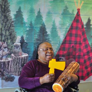 A cheerful woman in a wheelchair holds a toy axe and a drum, set against a vibrant forest-themed mural.