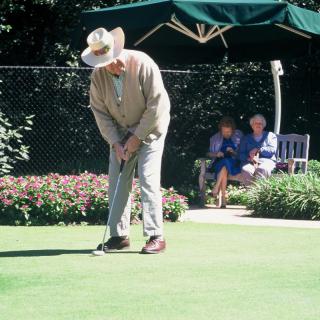 An elderly man putting on a golf course while others watch from a bench.