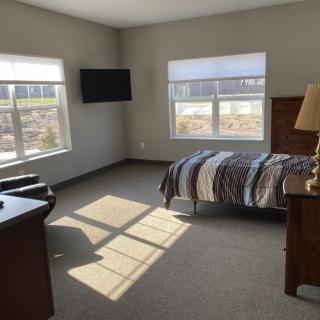 A well-lit bedroom with a single bed, a television mounted on the wall, and two large windows allowing natural sunlight to fill the space.
