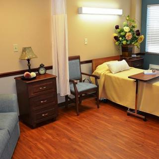 A well-appointed hospital room featuring a single bed, a chair, a bedside table with a lamp, and decorative flowers.