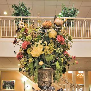 A large, ornate floral arrangement in a vase displayed indoors with plants and a staircase in the background.