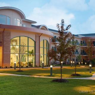 A modern building with large windows, surrounded by a well-lit garden and trees during the evening.