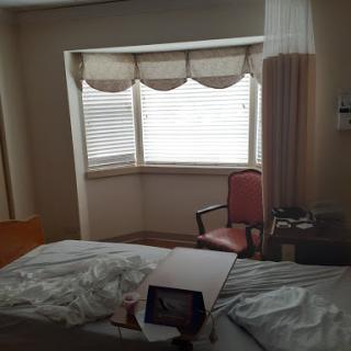 A view of a hospital room featuring a bed, a window with curtains, and a chair.