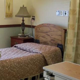 A hospital room with a neatly made bed, nightstand, lamp, and curtain.
