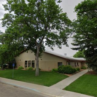 A single-story building surrounded by trees and a grassy lawn, with a sidewalk leading up to the entrance.