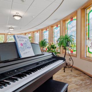 A bright music room with a piano, featuring stained glass windows and indoor plants.