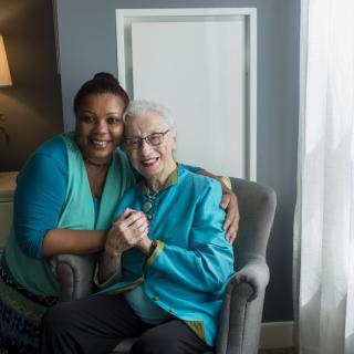 A caregiver and an elderly woman smiling warmly while sitting together in an armchair.