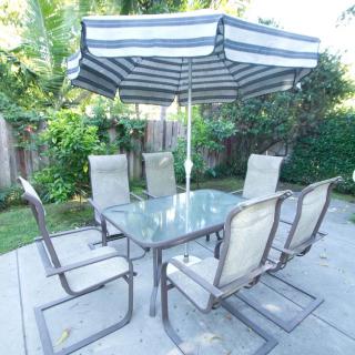 A glass patio table with six chairs and a striped umbrella, set in a garden.