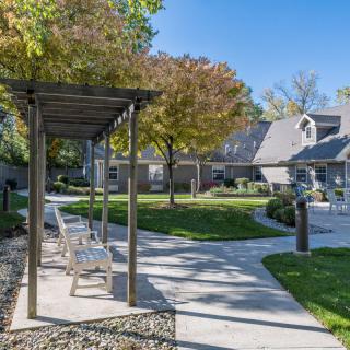 A peaceful garden pathway with benches under a pergola, surrounded by trees and houses on a sunny day.