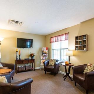 A cozy living room with comfortable chairs, a table, a mounted television, and a large window allowing natural light to enter.