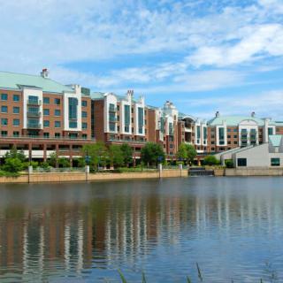 A modern building complex reflected in the calm waters of a river under a blue sky.