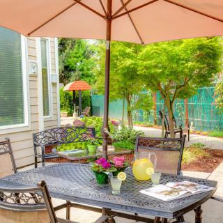 A cozy outdoor patio setting with a table, chairs, and an umbrella surrounded by lush greenery.