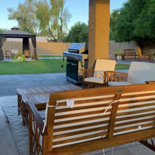 A cozy backyard patio with wooden furniture and a grill, overlooking a lawn and gazebo.