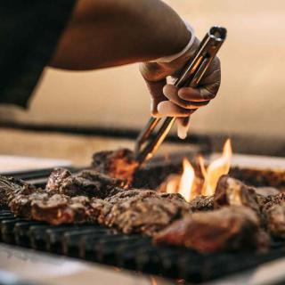A person grilling meat over an open flame using tongs.