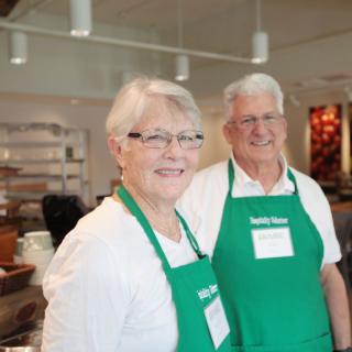A senior couple volunteering in a kitchen, wearing green aprons and smiling.