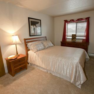 A cozy bedroom with a neatly made bed, nightstand, and lamp next to a window with red curtains.