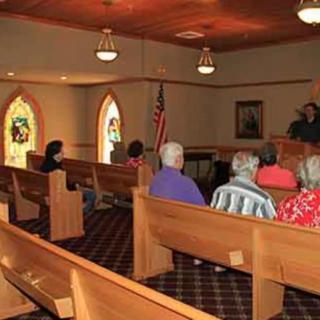A small congregation gathered inside a church with stained glass windows and wooden pews.