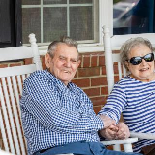 An elderly couple sitting on rocking chairs, smiling and holding hands.
