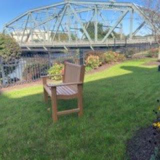 A peaceful outdoor scene featuring a wooden bench on green grass with a bridge over a river in the background.