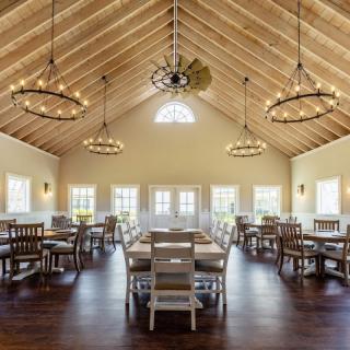 A spacious dining room with wooden ceilings and elegant chandeliers, featuring neatly arranged tables and chairs illuminated by natural light.