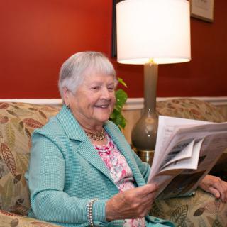 An elderly woman smiles while reading a newspaper in a cozy living room setting.