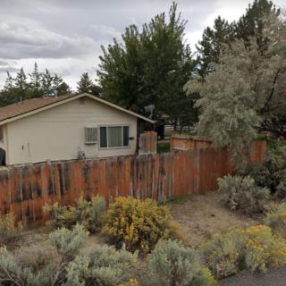 A suburban house with a wooden fence, surrounded by trees and bushes under an overcast sky.