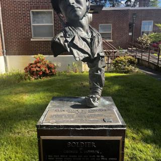 A striking bust sculpture commemorating a notable individual, set in a green outdoor space.
