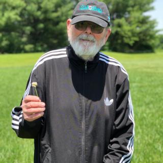 A bearded man in a black jacket and hat standing outside on a grassy field.
