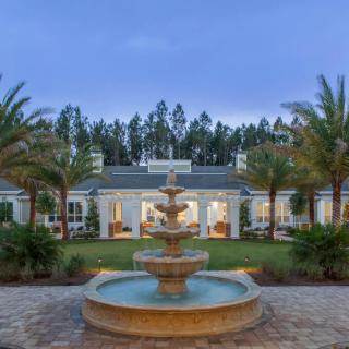 A luxurious mansion with a grand fountain surrounded by palm trees during the evening.