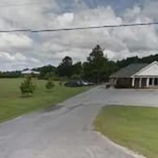 A small building with a driveway surrounded by grass and trees under a cloudy sky.