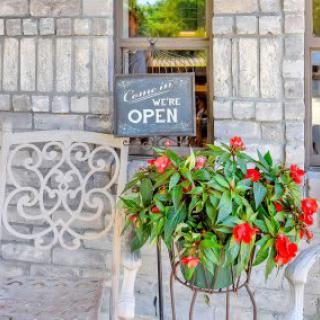 An inviting cafe entrance with a 'Come In, We're Open' sign surrounded by vibrant red flowers and a stone wall backdrop.