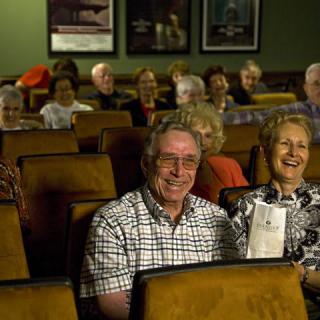 A group of seniors enjoying a movie at the cinema, with two people prominently laughing and holding popcorn.