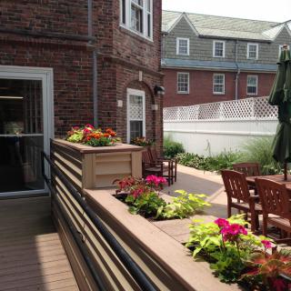 A sunny patio area with colorful flowers, garden furniture, and a brick building in the background.