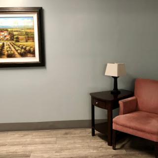 A cozy lounge area featuring a pink chair, a small table with a lamp, and framed artwork on the wall.
