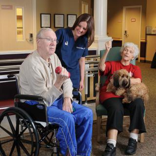 A nurse interacts with an elderly man in a wheelchair and a woman holding a dog in a care home setting.