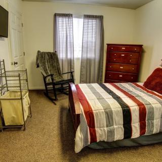 A cozy bedroom with a bed, dresser, rocking chair, and a wall-mounted television.