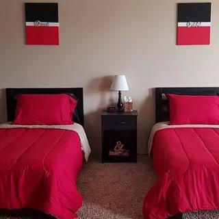 A neatly arranged bedroom with two twin beds featuring red bedding, separated by a nightstand with a lamp and wall art above each bed.