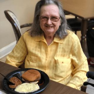 An elderly woman smiling while sitting in front of a meal at a table.