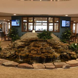 An indoor lobby featuring a central stone fountain surrounded by plants and decorative elements.