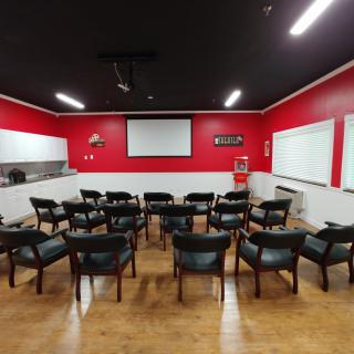 A classroom or meeting room with rows of chairs facing a projector screen, set against red walls.