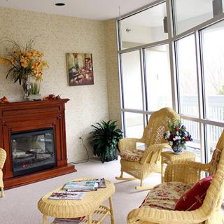 A cozy living room with a fireplace and wicker furniture, featuring large windows and decorative plants.