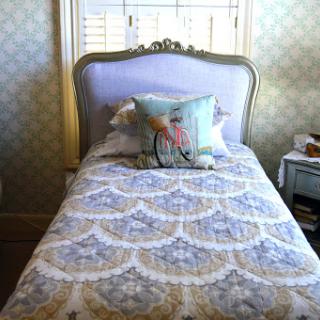 A neatly made twin bed with decorative bedding and a charming pillow, set against a patterned wallpaper in a cozy bedroom.