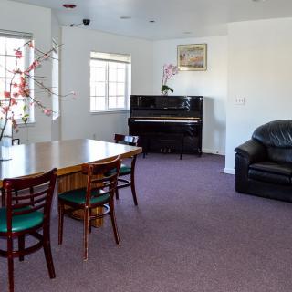 A cozy living room featuring a piano, dining table with chairs, a couch, and large windows.